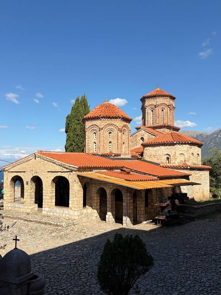 Stone monastery complex with red-tiled domes and arches set against blue skies and distant hills.