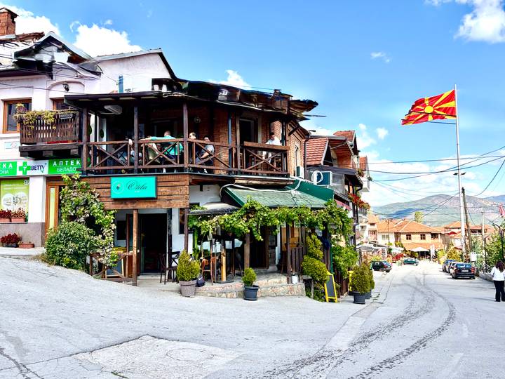 Wood-framed café on a hillside street flying the Macedonian flag under a bright blue sky.