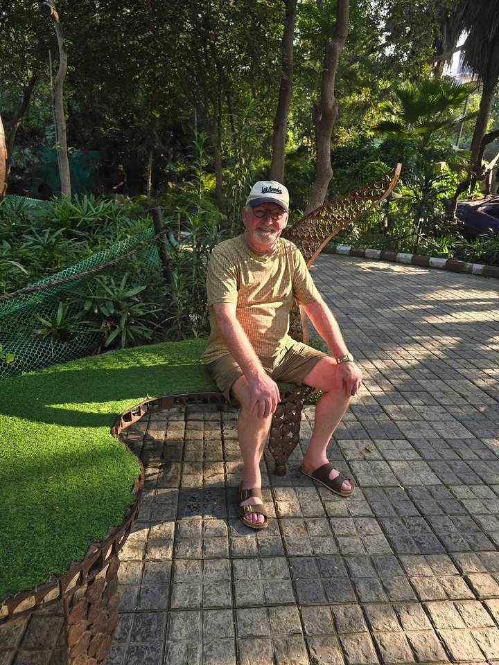 Older man sitting on a leaf-shaped bench in a sunny garden path.