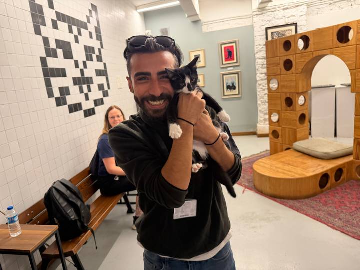 Man happily holds a black-and-white kitten inside a modern cat café while a traveler smiles in the background.