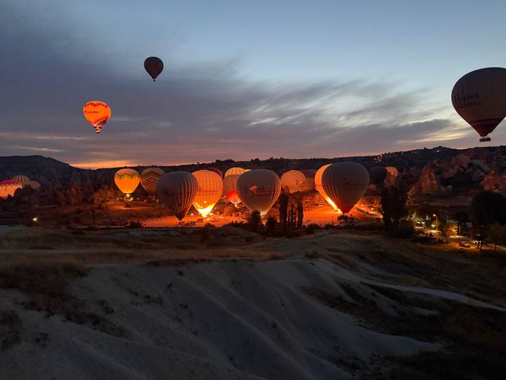 Pre-dawn scene of tethered hot air balloons glowing orange as their burners ignite against a purple sky.