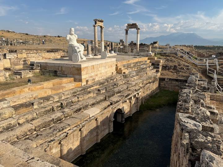 Ancient Greco-Roman ruins featuring a seated marble statue overlooking stepped stone pools with distant mountains.