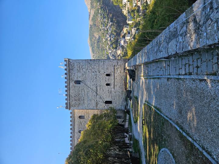 Stone castle wall and path overlook valley town under clear blue sky.