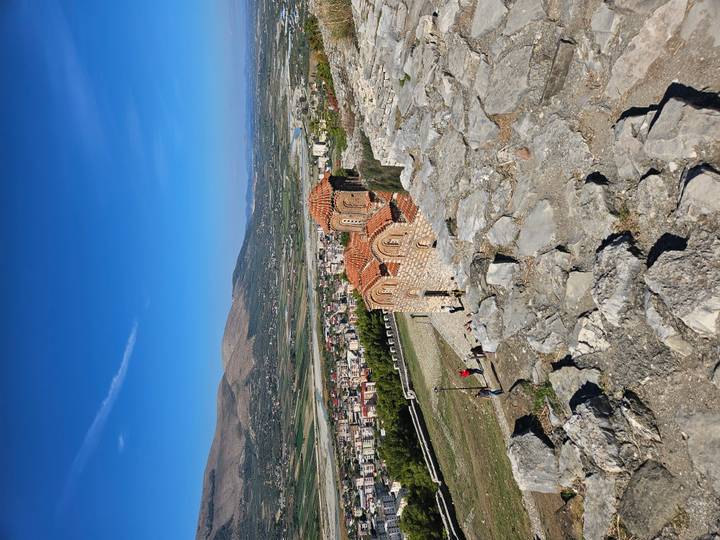 Small hillside Byzantine church overlooks broad Albanian valley with tourists nearby.