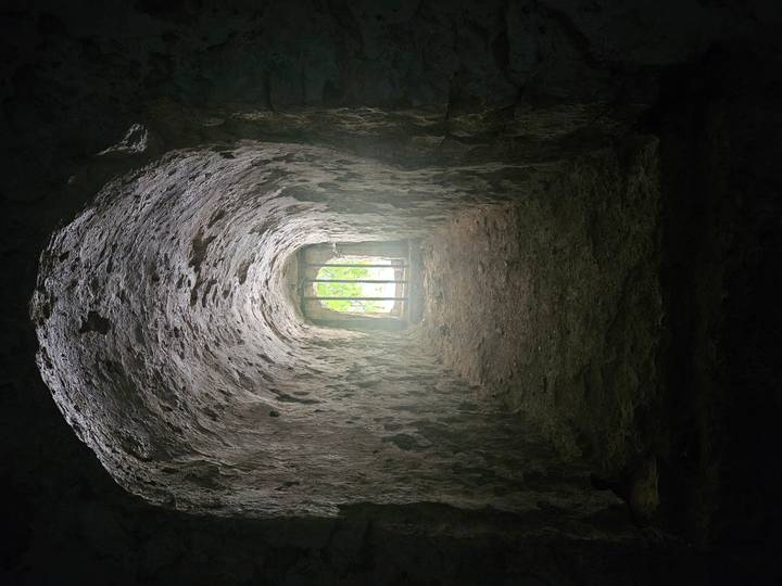 Rough stone tunnel interior leads to barred window with greenery beyond.