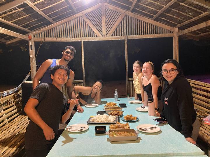 Friends sit around a long table of local dishes in a rustic beach hut at night.