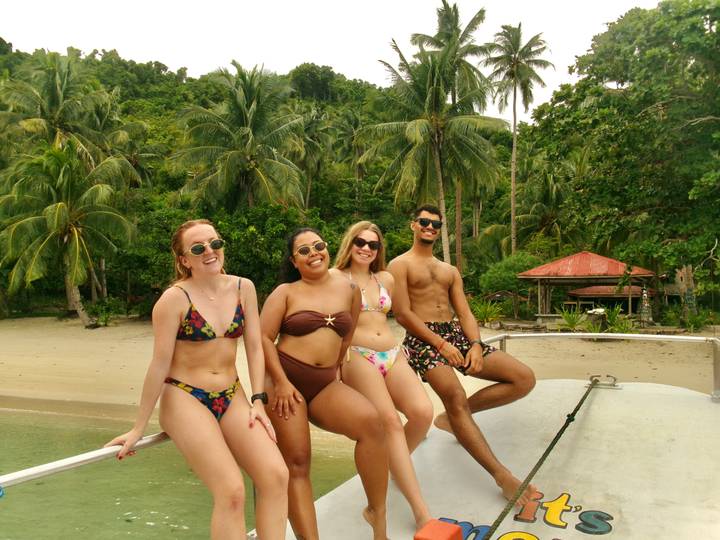 Four travellers in swimwear pose on a boat with palm-fringed beach and jungle backdrop.