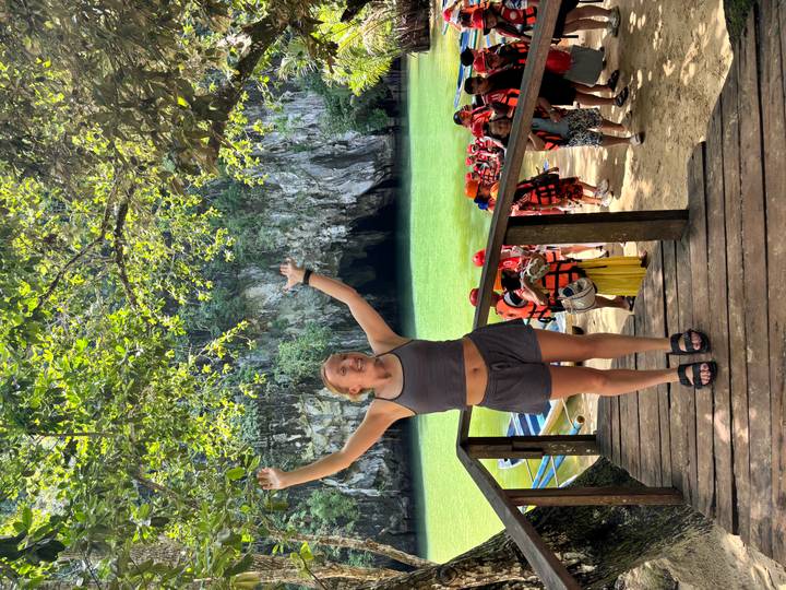 A traveller raises her arms on a wooden platform overlooking emerald water and a cave entrance crowded with tour boats.