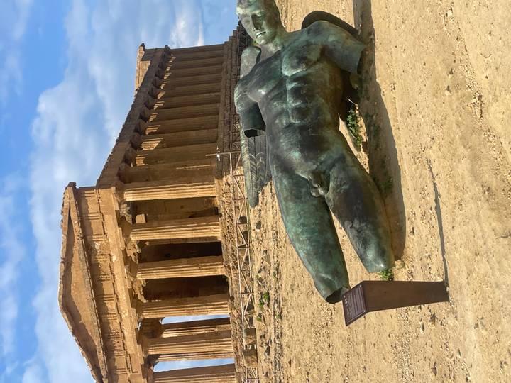 Ancient Doric temple ruin with modern bronze fallen statue in foreground on sandy terrain.
