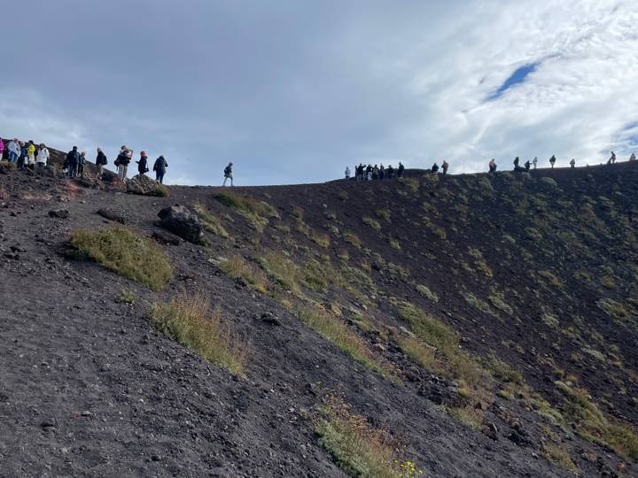 Hikers walking along the rim of a volcanic crater with dark lava rocks and sparse vegetation.