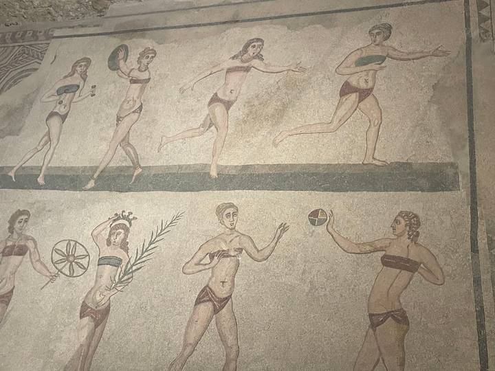 Ancient Roman mosaic of women playing sports wearing bikinis on tan background.