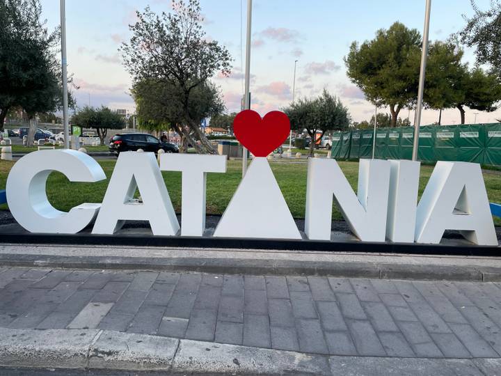 Large white and red love-themed CATANIA sign near a city street with trees behind.