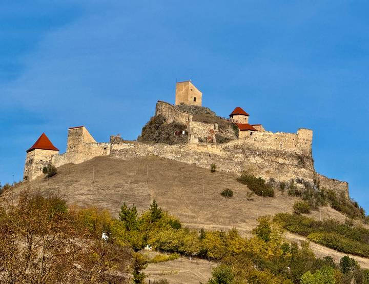 Hilltop medieval fortress ruins with tan stone walls and red roofs under blue sky.