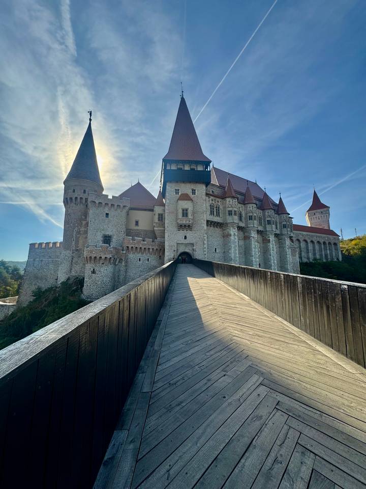 Stone Gothic-style castle with red roofs approached by a long wooden bridge on a sunny day