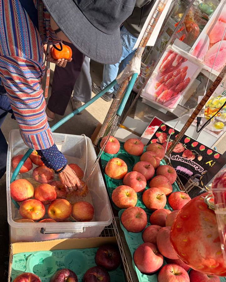 Market stall with fresh apples being washed in a plastic tub and fruit displays all around.