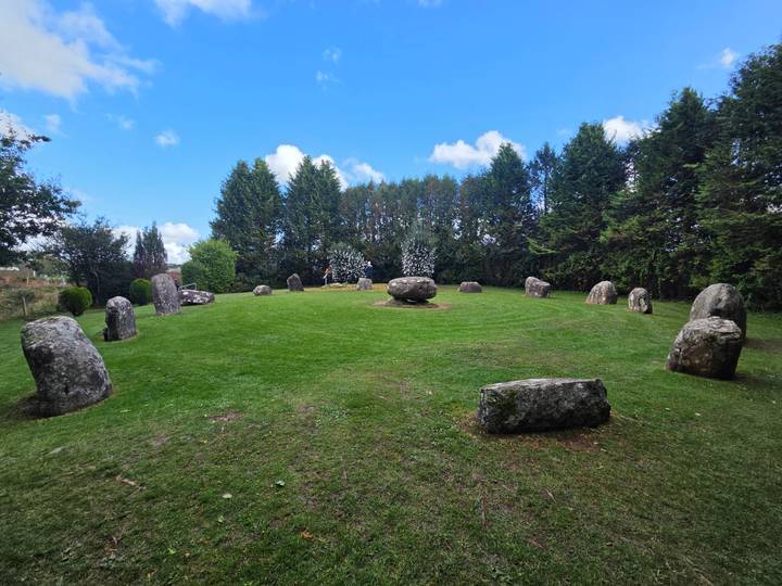 Grassy stone circle with weathered boulders arranged on a manicured lawn under blue sky.