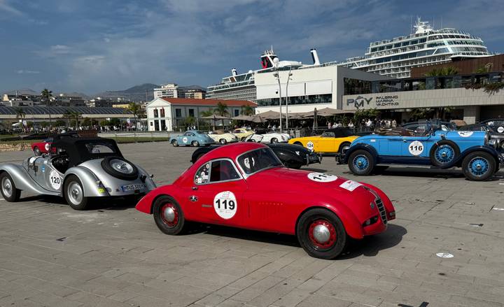 Classic sports cars displayed on a waterfront plaza with a docked cruise ship towering behind