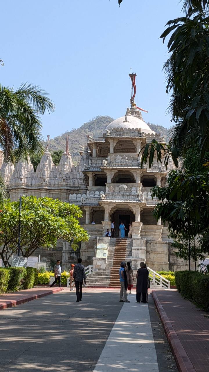 Intricately carved white marble Jain temple complex set against a hillside with visitors at the entrance.