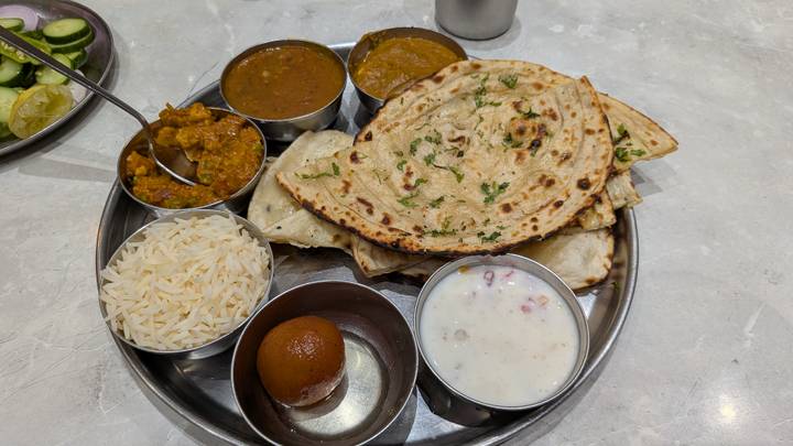 Traditional Indian thali featuring assorted curries, rice, flatbreads, raita and a gulab jamun dessert served on a metal platter.
