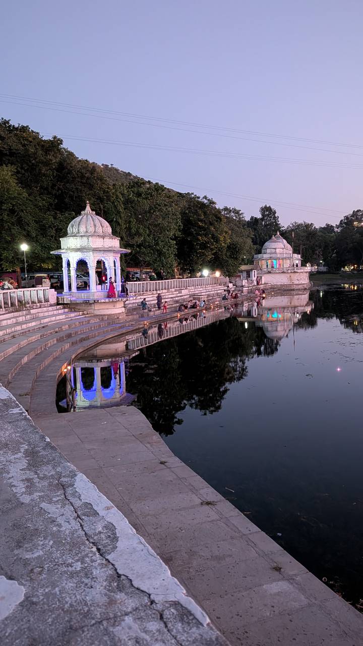 Evening scene of lakeside ghats with small temples lit by colorful lights reflecting on still water while people gather along the steps.
