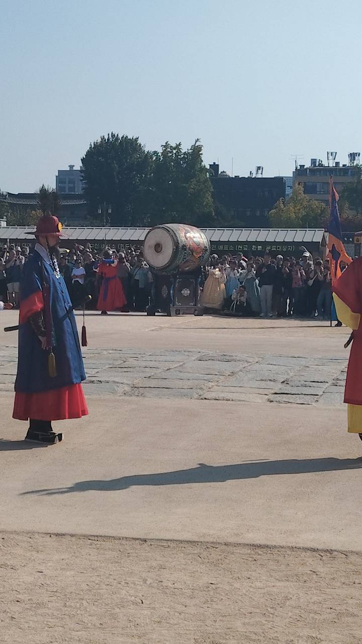 Traditional guard ceremony at a Korean palace courtyard with colorful robes, drum and onlooking crowd.