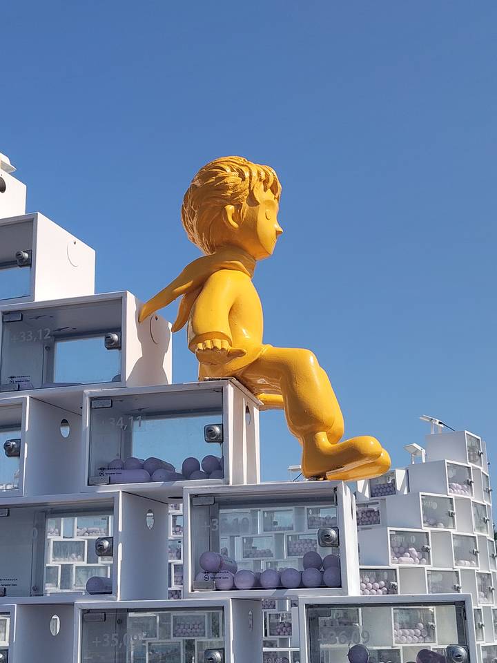 Bright yellow Little Prince statue perched on a modern white structure against a clear blue sky.