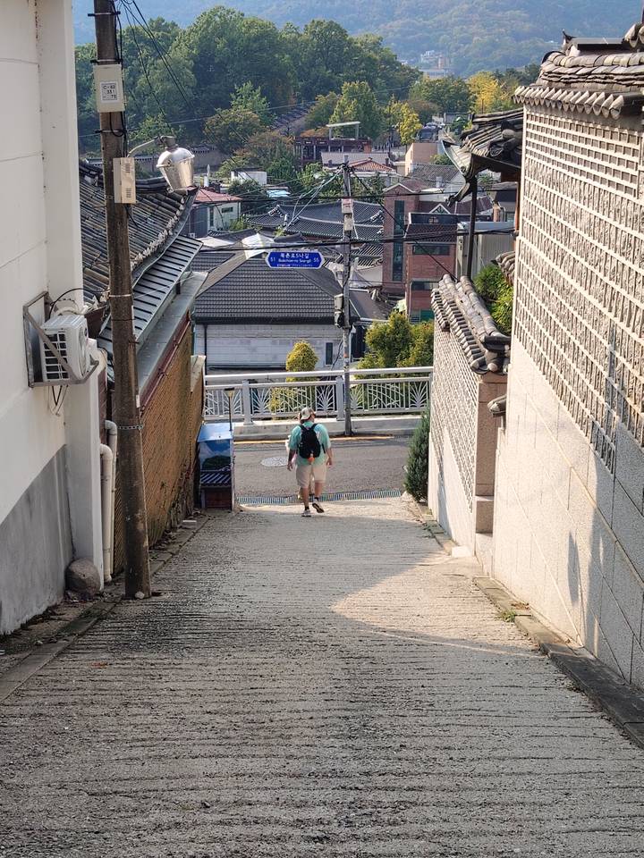 Lone traveler with backpack walking up a narrow stone alley between traditional Korean walls.