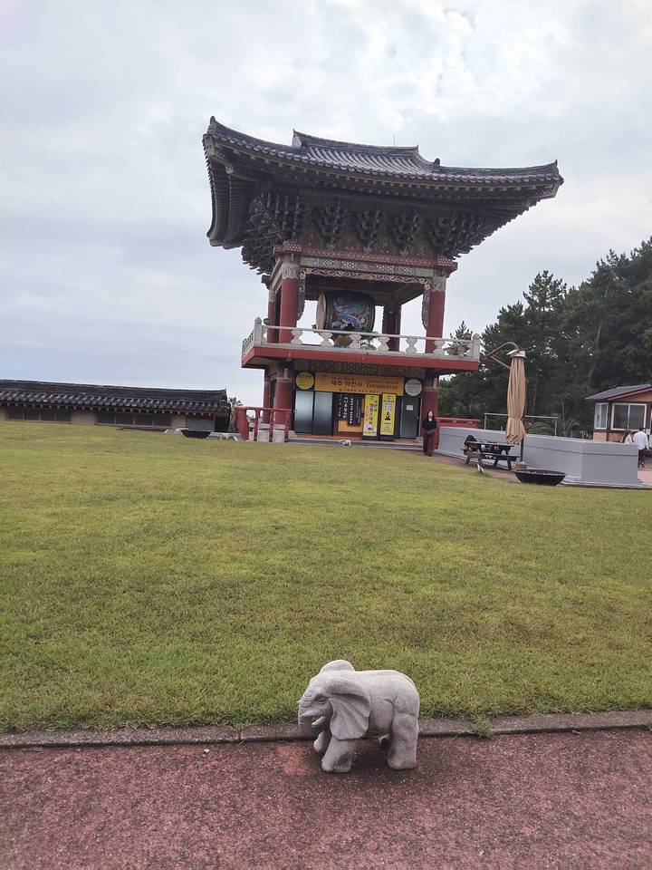 Wide grassy lawn leading to a colorful Buddhist bell tower pavilion on an overcast day.