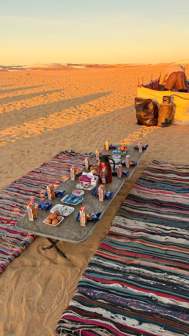 Outdoor breakfast spread on rugs and low tables laid out on desert sand at dawn.