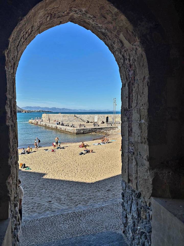 View through a stone arch to a sandy town beach with sunbathers and a stone pier jutting into turquoise water.