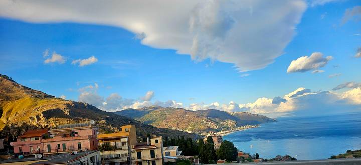 Panoramic coastal town with colourful houses and rolling hills meeting a deep blue sea beneath dramatic clouds.