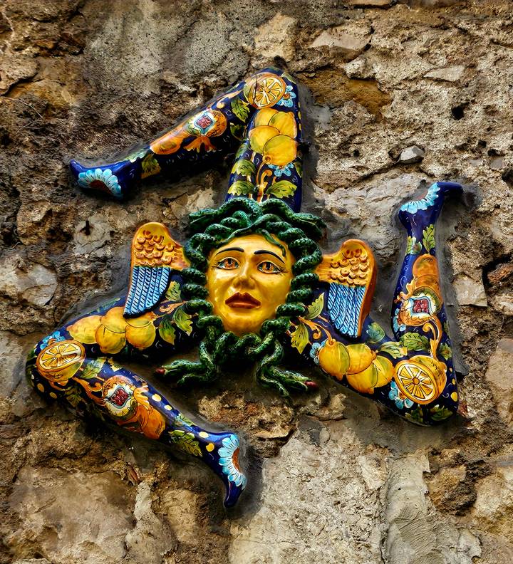 Colourful ceramic Trinacria symbol with Medusa head and Sicilian motifs mounted on stone wall.