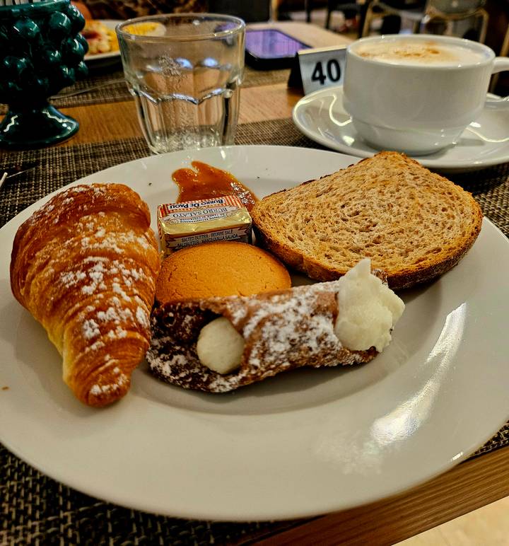 Breakfast plate with croissant, cannoli, biscuit, jam and slice of bread on white dish.