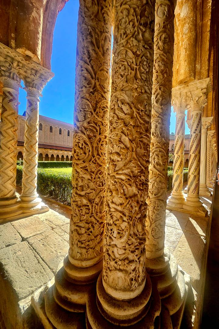 Sunlit close-up of elaborately carved stone column and cloister arches.