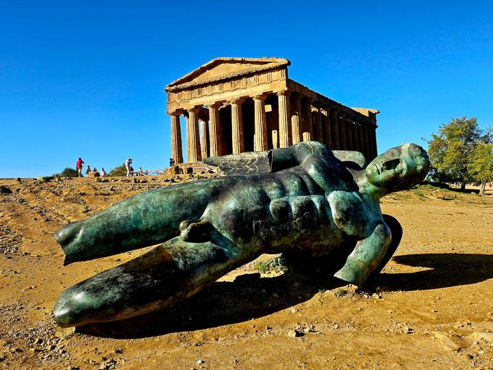 Bronze fallen Icarus statue lying before the well-preserved Temple of Concordia at the Valley of the Temples on a sunny day.