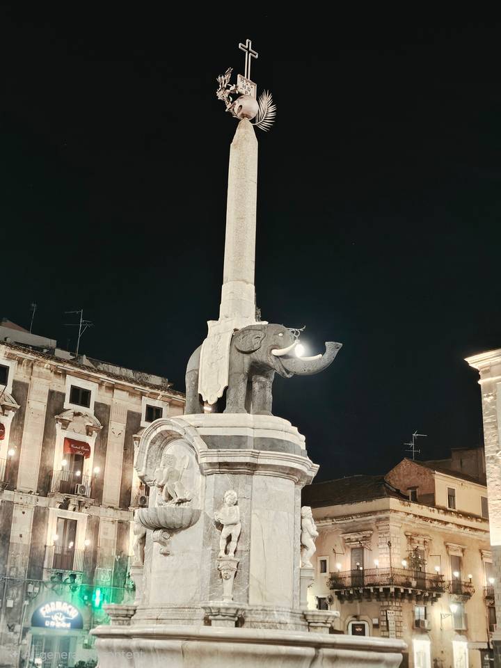 Night view of Catania’s elephant fountain topped obelisk illuminated in a city square.