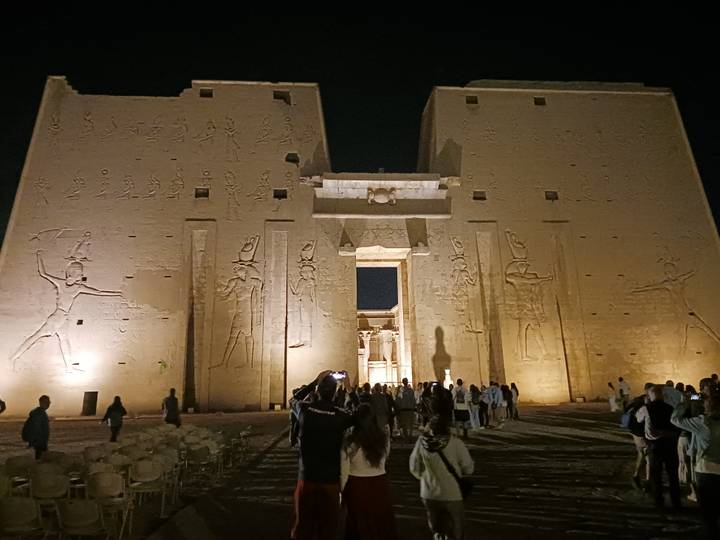 Night tourists entering Edfu Temple façade lit with warm lights revealing carved hieroglyphs.