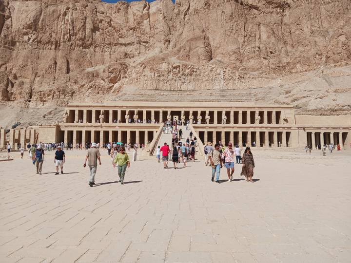 Wide courtyard view of the terraced Temple of Hatshepsut set against rugged cliffs with tourists approaching.