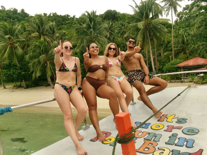 Four young travellers in swimsuits posing on the bow of a boat near a tropical beach.