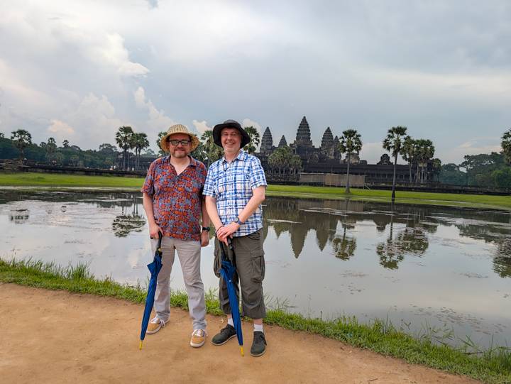 Two tourists stand with umbrellas by the reflective pond opposite Angkor Wat’s iconic towers