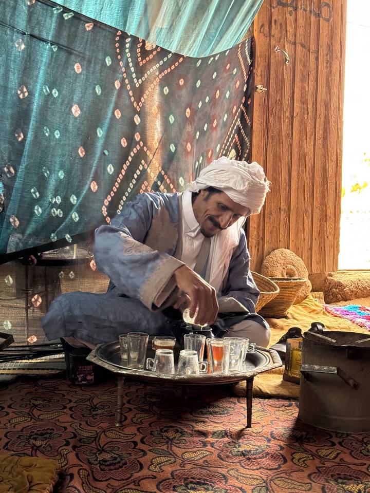 Berber man in traditional attire pours mint tea inside a tent decorated with textiles.