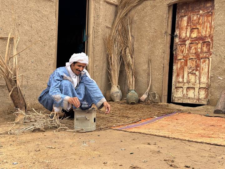 Berber man squats outside an adobe home brewing tea on a small metal stove.