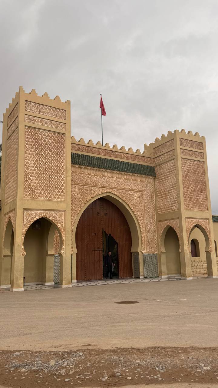 An ornate Moroccan city gate with intricate geometric carvings and pointed arches under an overcast sky.