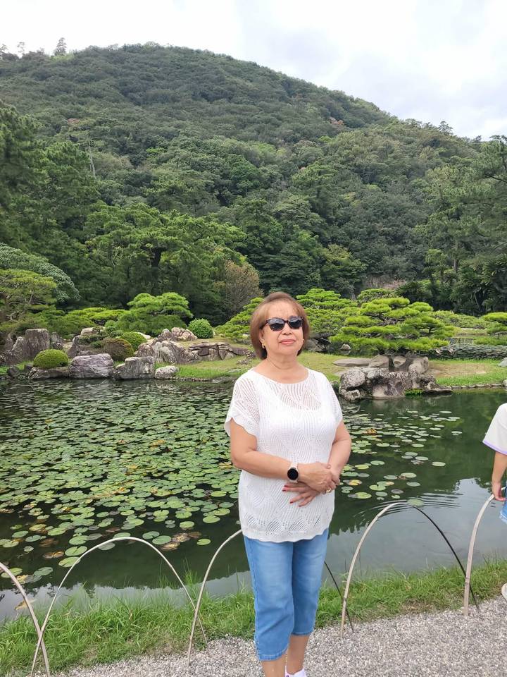 Woman stands beside a lily-covered pond in a tranquil Japanese garden surrounded by green trees.