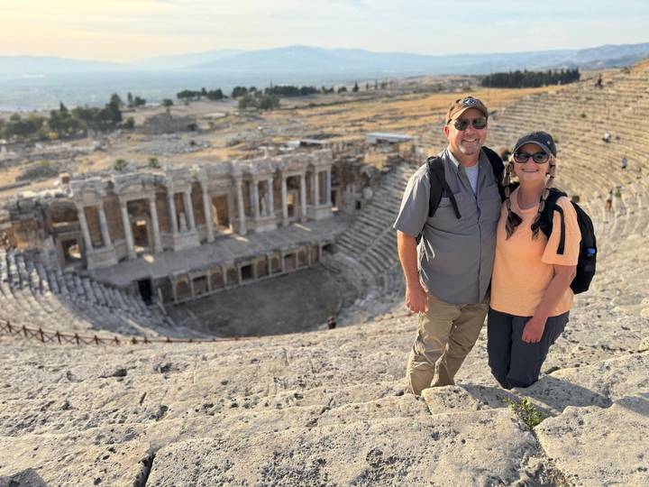 A smiling couple stands on the stone seating of the ancient Hierapolis theater overlooking the stage.