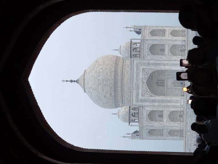 Crowds photograph the Taj Mahal framed by a dark archway in early morning haze.