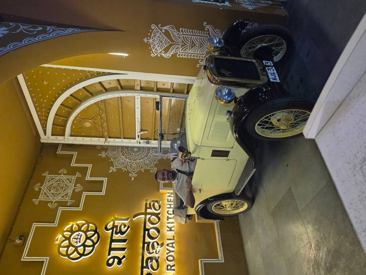 A smiling man poses in a vintage open-top car inside a stylishly decorated restaurant entrance.