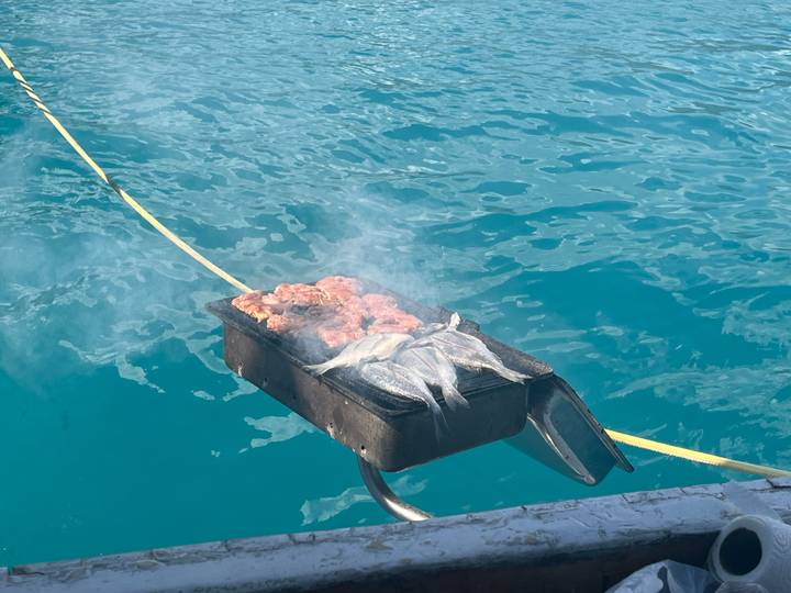 Small grill loaded with fish and meat smoking over bright turquoise sea water, likely on a boat.