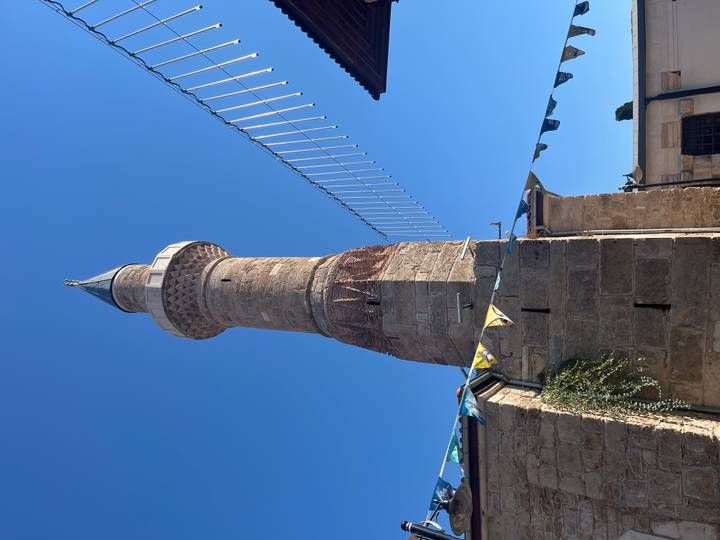 Stone minaret of a historic mosque rising against a clear blue sky with festive bunting below.