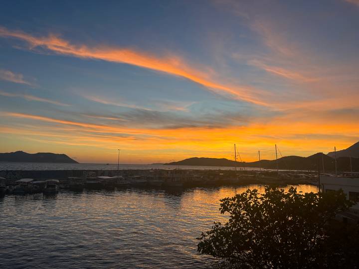 Colorful sunset over a marina with yachts and distant hills reflecting on calm water.